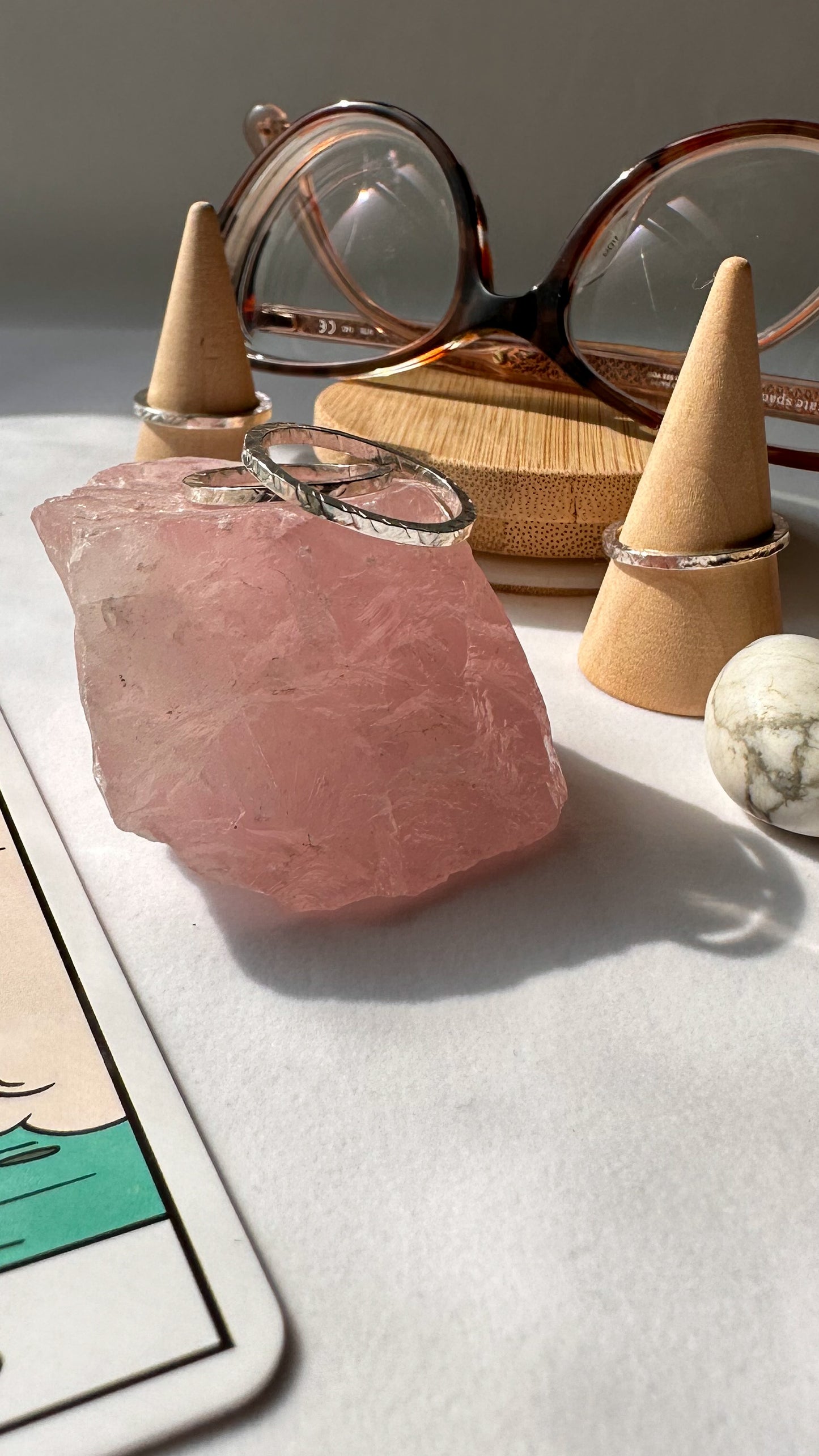 Two hammered texture silver rings are entwined on top of a piece of rose quartz. In the background two more silver hammered rings are on wooden cones. A pair of eyeglasses, a small howlite stone and the corner of a tarot card is visible.