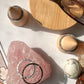 Photo taken from above of two entwined silver hammered texture rings on top of a chunk of rose quartz. Two more hammered rings are on top of wooden cones and a pair of eyeglasses sit on a small circle of wood. Small crystals and the corner of a jeweler's hammer are visible.