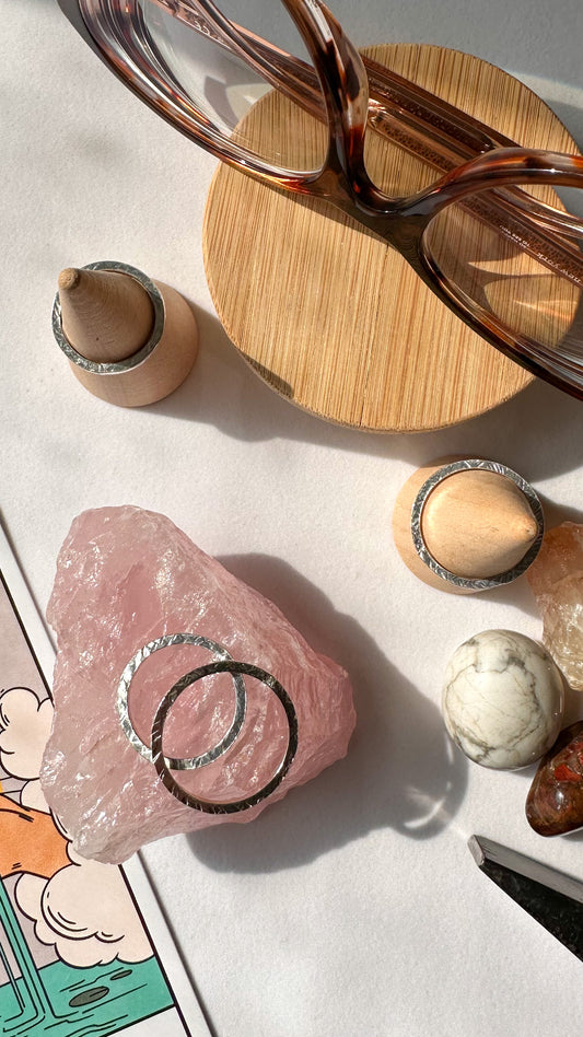 Photo taken from above of two entwined silver hammered texture rings on top of a chunk of rose quartz. Two more hammered rings are on top of wooden cones and a pair of eyeglasses sit on a small circle of wood. Small crystals and the corner of a jeweler's hammer are visible.