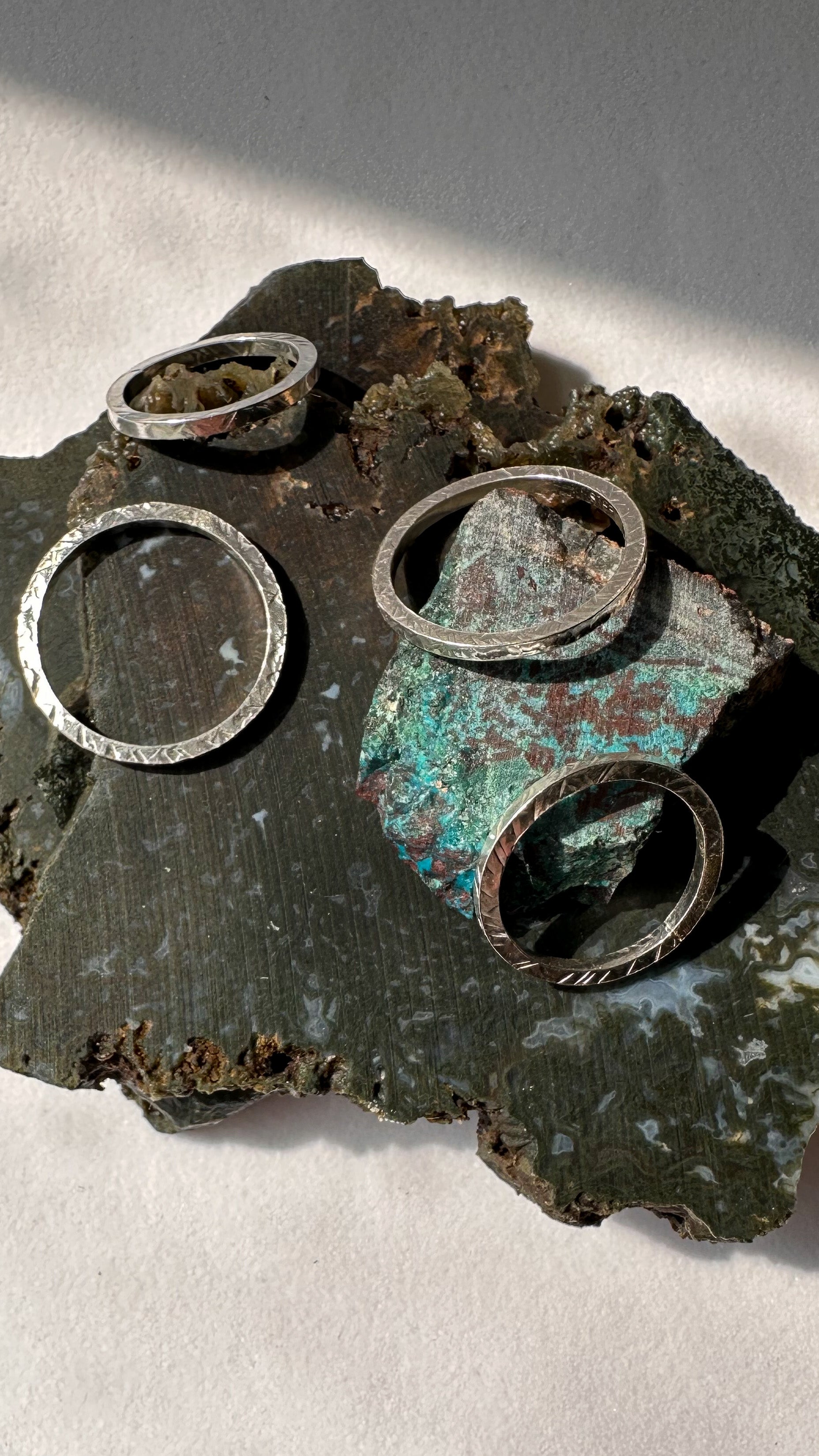 Four silver rings with hammered texture. They are arranged on two green slabs of moss agate and a small chunk of chrysocolla.